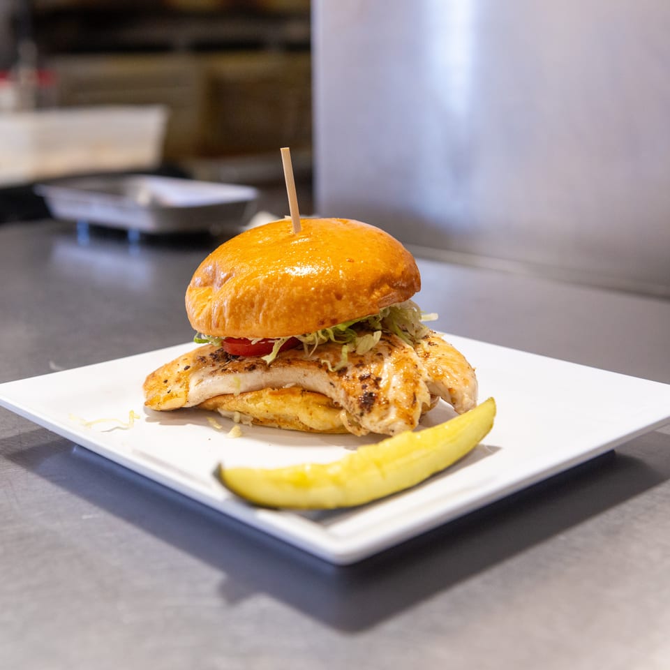 Grilled chicken sandwich with lettuce, tomato, and a pickle spear on a white plate.