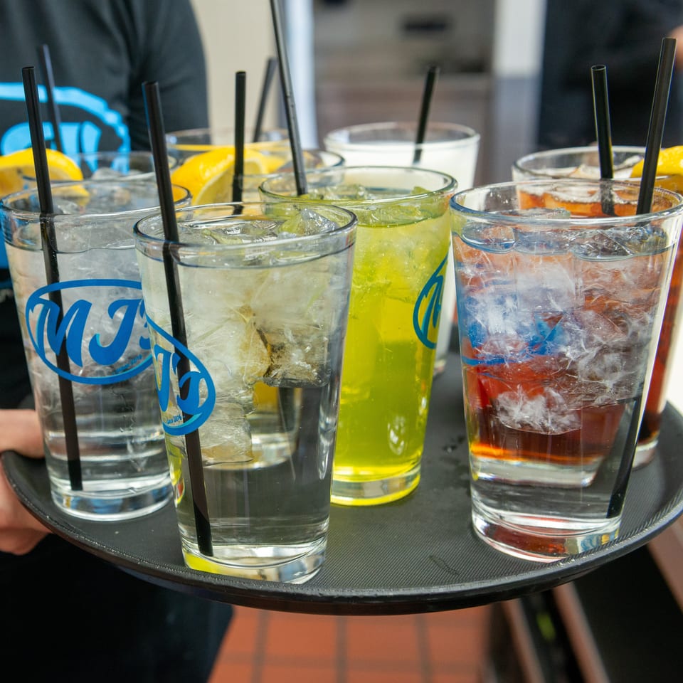 A server carries a tray of various iced drinks with straws and lemon garnishes.