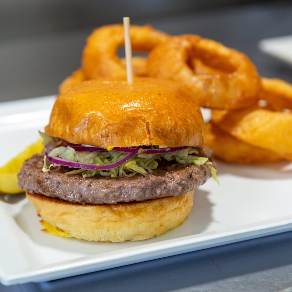 A burger with lettuce, red onion, and a pickle spear, served with onion rings on a white plate.