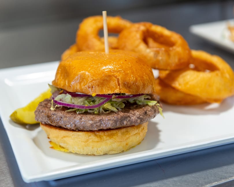 A burger with lettuce, red onion, and a pickle spear, served with onion rings on a white plate.